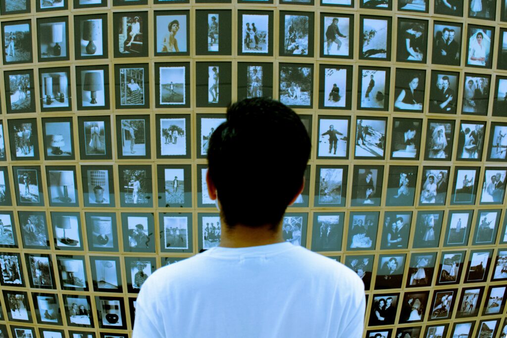 A collage of deepfake images that could mislead or deceive Person seen from behind wearing a white shirt, looking at a wall of small framed photos or polaroids arranged in a grid.